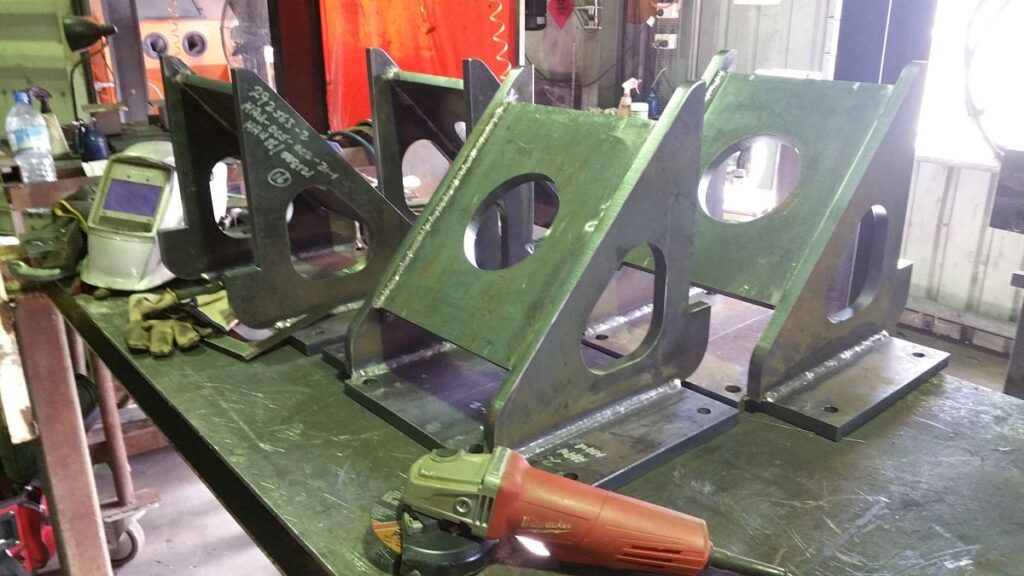 A workshop table with metal brackets featuring circular cutouts is ready for action. A red handheld grinder and welding tools, perfect companions for any project, sit nearby. The background showcases industrial equipment and a red curtain, while chalk markings suggest precise planning—contact Gorilla Weld for more.