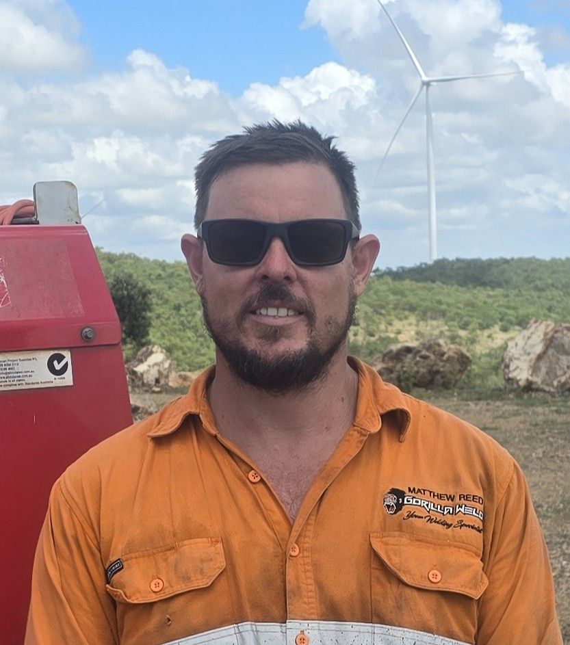A man wearing sunglasses and an orange work shirt stands outdoors. Behind him, a wind turbine and a hilly landscape with greenery are visible under a cloudy sky.