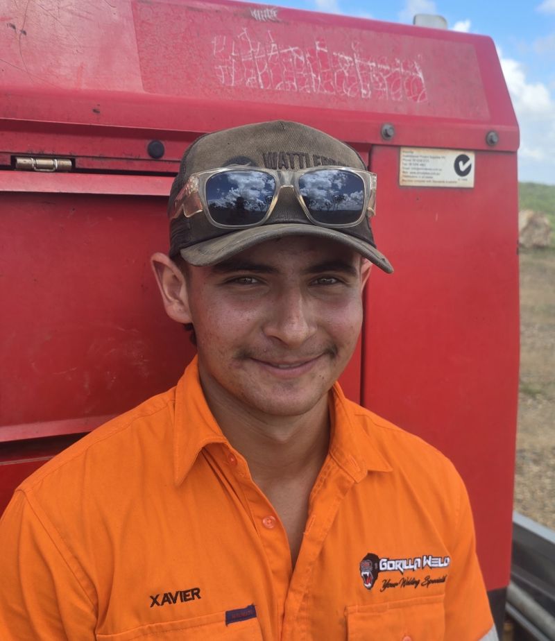 A person wearing an orange shirt with text, a cap, and sunglasses is smiling. They are standing in front of red machinery outdoors, with a partly cloudy sky in the background.